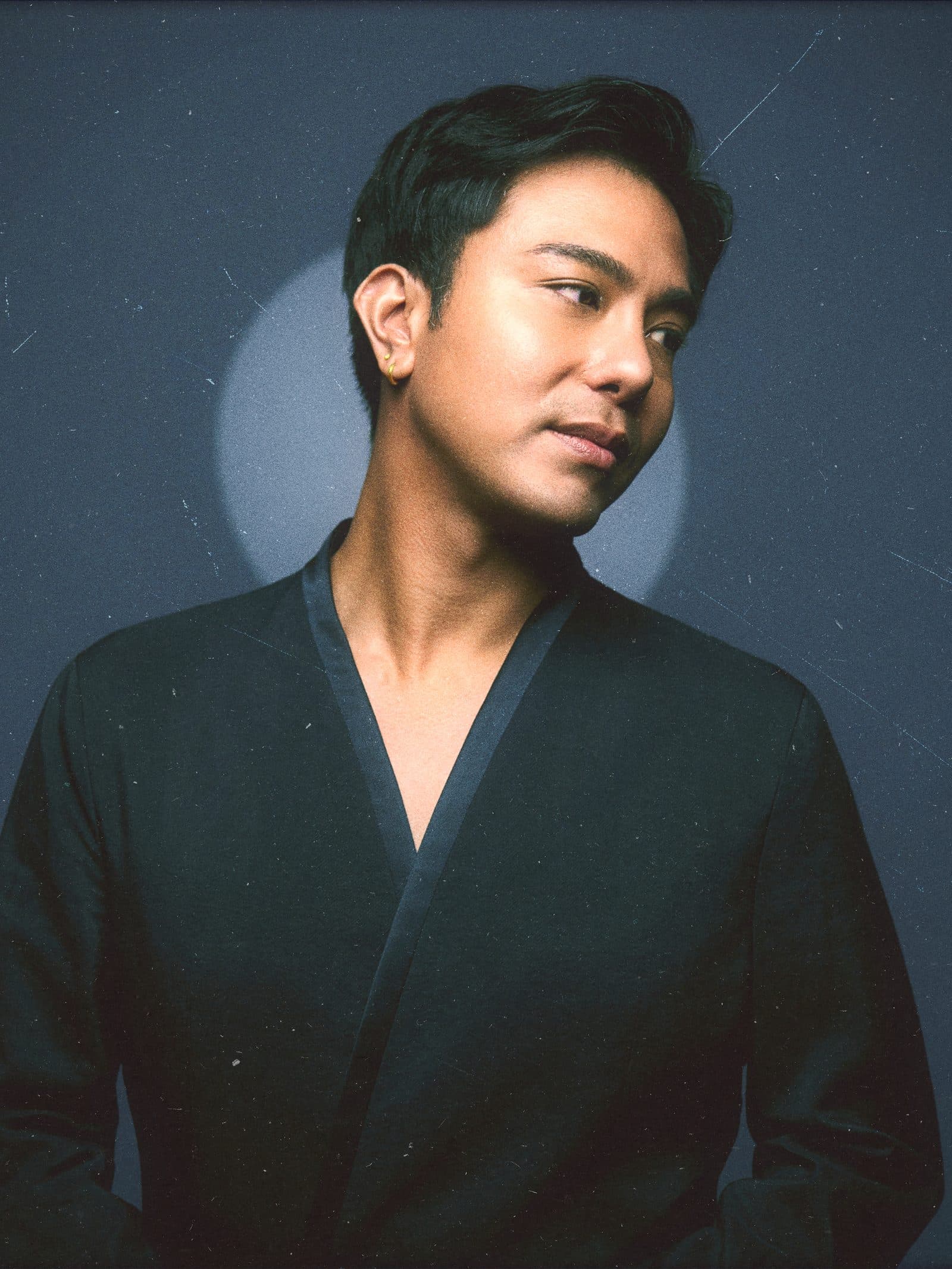 Raffy Bleu in a dark kimono jacket, turned from a single studio light against a navy backdrop
